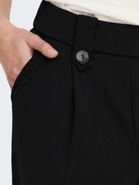Pantalon noir avec une poche latérale, fermé par un bouton de couleur foncée, confectionné dans un tissu lisse et texturé.