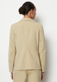 Blazer beige su misura con una texture liscia, maniche lunghe e schiena con un solo spacchetto. Linee pulite e design strutturato. Stile minimalista.