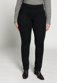 Schwarze Stretchhose mit hohem Bund, seitlichen Taschen, glatter Textur und schmal zulaufendem Bein, kombiniert mit schwarzen Stiefeln mit Absatz.