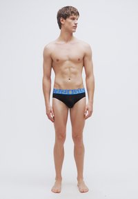 Μαύρα ανδρικά μαγιό τύπου σλιπ με μπλε σχέδιο ελληνικού κλειδιού στη μέση, διακοσμημένα με χρυσό έμβλημα. Το ύφασμα φαίνεται λείο και εφαρμοστό, σχεδιασμένο για άνεση.