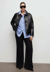 Black leather jacket with a collar, buttoned front, worn over a blue striped shirt. Black wide-leg pants and oversized sunglasses complete the look.