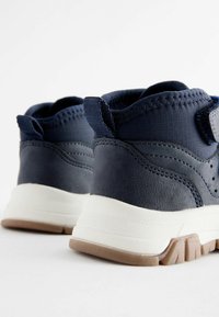 Mörkblå hög-top sneakers har en texturerad överdel i läder och tyg, vit mellansula och en brun gummisula med greppdetaljer.