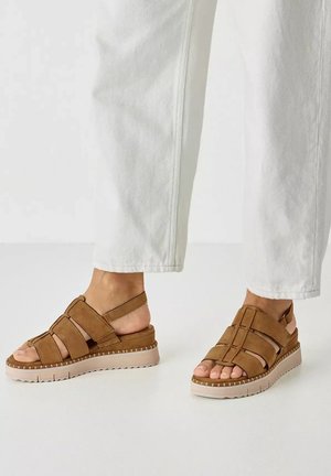 Füße in braunen Wildleder-Riemchensandalen mit dicken weißen Sohlen, kombiniert mit weißen verkürzten Hosen vor neutralem Hintergrund.