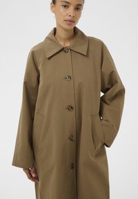 Femme portant un long manteau marron boutonné avec des poches, debout devant un fond clair uni.