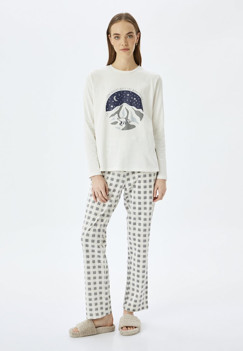 Top de manga larga color crema con un gráfico de montaña, combinado con pantalones de cuadros blancos y grises y zapatillas beige de felpa.