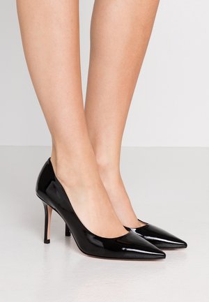 Jambes portant des escarpins noirs brillants à bout pointu avec talons aiguilles fins sur un sol et un fond clairs.