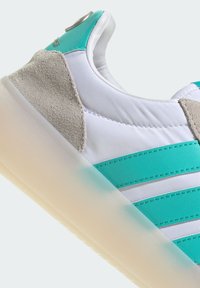 White sneaker with turquoise accents, suede heel, and three stripes. Features a translucent sole and stitched detailing.