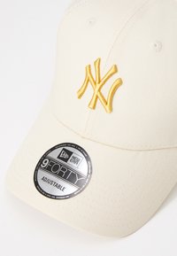 Beige kasket med gult broderet NY-logo. Har en rund skygge, tekstureret stof, justerbar rem og et rundt mærke med betegnelsen "9FORTY."