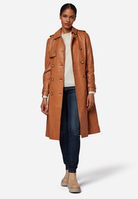 Brown leather trench coat with double-breasted buttons, belt detail, and shoulder epaulets, paired with a light sweater and dark jeans.