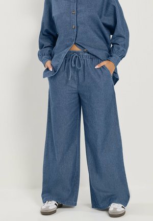 Loszittende blauwe spijkerbroek met een trekkoord in de taille en zijzakken, gedragen met een bijpassend blauw doorgestikt overhemd en witte sneakers.