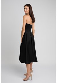 Czarna, bezramienna sukienka midi z dopasowanym gorsetem i rozkloszowanym dołem, wykonana z gładkiego materiału. Noszona z beżowymi sandałami na wysokim obcasie.