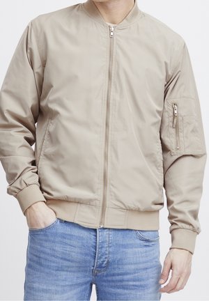 Beige Bomberjacke mit durchgehendem Reißverschluss, mit Reißverschluss versehener Tasche am linken Ärmel, gerippten Bündchen und Saum, getragen mit hellblauen Jeans.