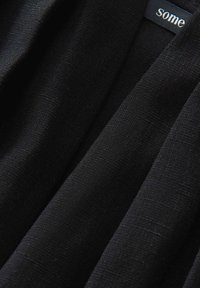 Tissu noir avec une surface lisse et légèrement texturée, plié pour montrer la profondeur et le drapé. Étiquette visible avec le mot "some" en blanc.