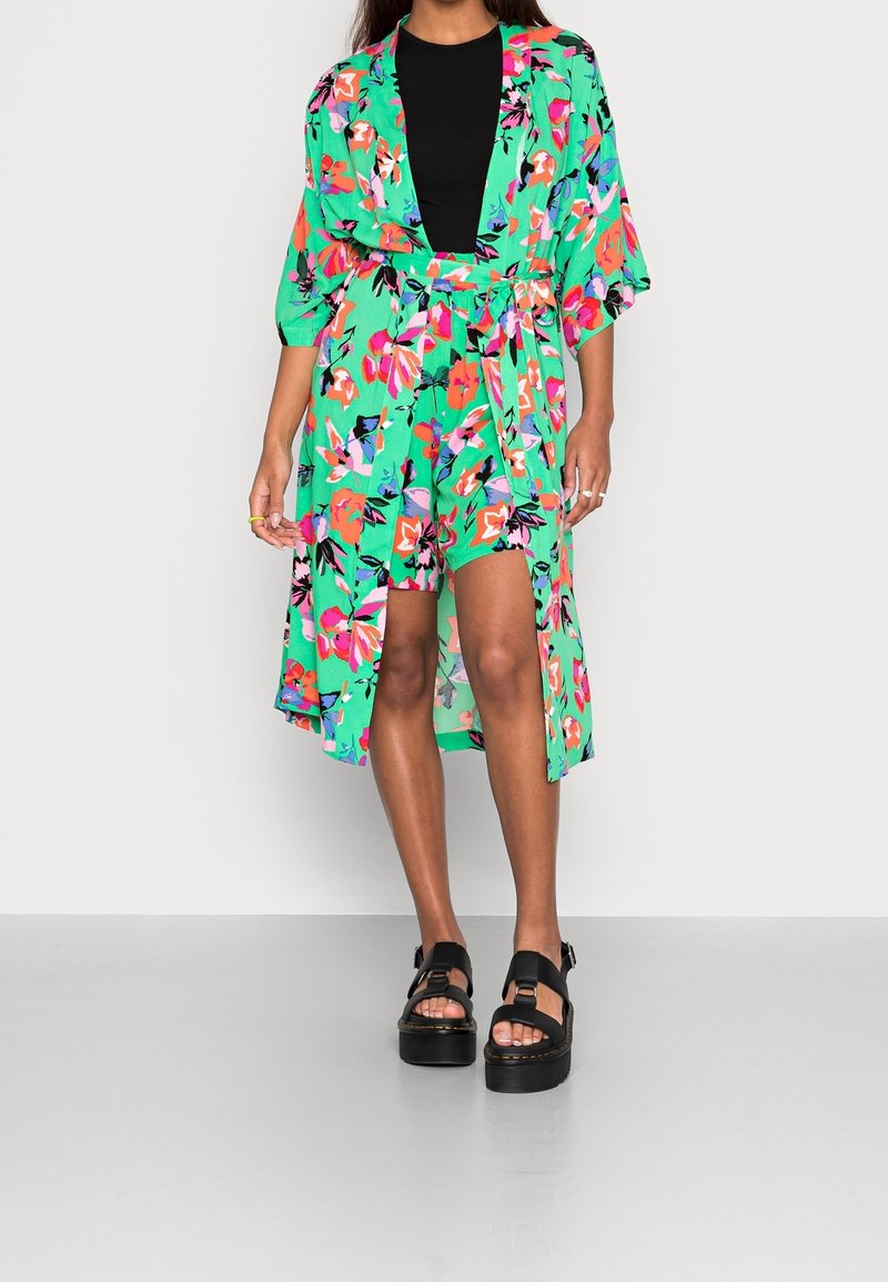 Femme portant une robe portefeuille verte à fleurs avec de larges manches sur un top noir, associée à des sandales plateformes noires, debout sur un sol gris.