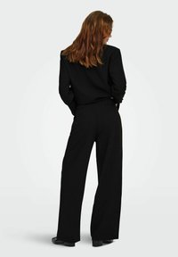 Veste de costume noire sur mesure et pantalons à jambe large en tissu texturé, présentant une coupe classique et une silhouette perdue. Chaussures non visibles.