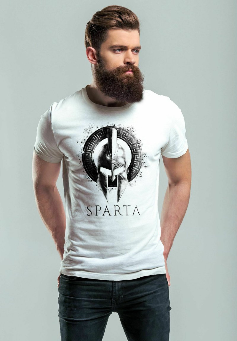 White t-shirt featuring a black spartan helmet graphic with the text "SPARTA" below. Model is wearing dark jeans and has a beard.