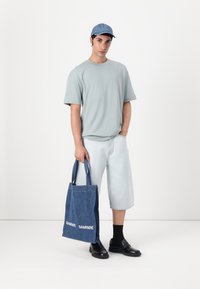 Denim torba s plavim trakama, bijelim logotipom, svijetloplava majica, svijetle kratke hlače i crne mokasine na osobi koja stoji protiv jednostavne pozadine.