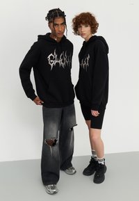 Zwarte hoodies met een zilveren grafisch logo, een voorzak en geribbelde manchetten. Het model links draagt versleten grijze jeans; het model rechts heeft zwarte shorts aan.