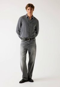 Sivá zapínacia košeľa a vybledlé sivé rifle, doplnené čiernym opaskom a zladenými topánkami. Oblečenie má ležérny, moderný dizajn.