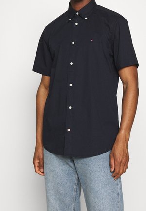 Man wearing black short-sleeve button-up shirt with small logo on chest, paired with light blue jeans against white background.