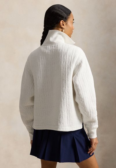 Λευκό καπιτονέ πουλόβερ με ψηλό γιακά και άνετη εφαρμογή. Διαθέτει ριμπ μανσέτες και τελείωμα, συνδυασμένο με μπλε σκούρα πλισέ φούστα.