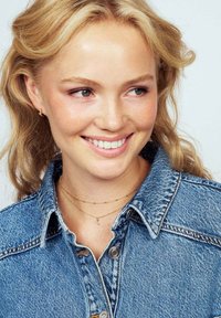 Leende ung kvinna med vågigt blont hår som bär en blå jeansjacka och lager av guldhalsband mot en ljus bakgrund.