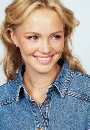 Leende ung kvinna med vågigt blont hår som bär en blå jeansjacka och lager av guldhalsband mot en ljus bakgrund.