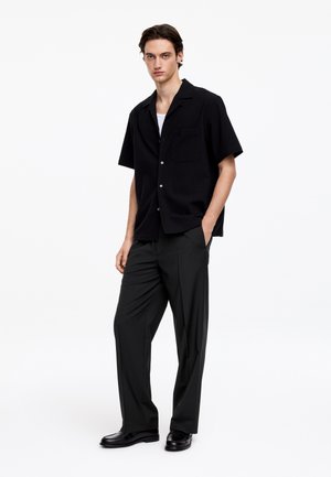 Giovane uomo in piedi con le mani in tasca che indossa una camicia nera a maniche corte, pantaloni neri, scarpe nere e una canottiera bianca su sfondo bianco.