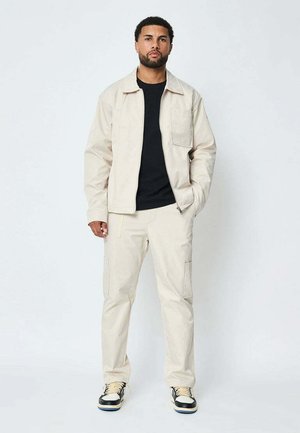 Homme barbu portant une veste beige, un pantalon cargo assorti, une chemise noire et des baskets noir et blanc, debout devant un fond blanc uni.