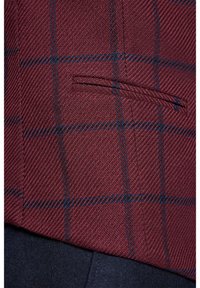 Tkanina w burgundową kratę z niebieskimi liniami; posiada teksturowane sploty oraz kieszeń z lamówką. Dół wykonany z ciemnego materiału, tworzącego kontrast.