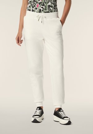 Pantalons de survêtement en coton blanc avec une taille à cordon, des jambes effilées et une broderie de marque sur la hanche gauche, associés à des baskets noires et blanches.