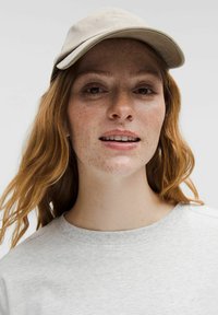 Beige kasket med buet skygge, logo på forsiden, båret med en lys grå sweatshirt. Håret er bølget og rødbrunt med synlige fregner.