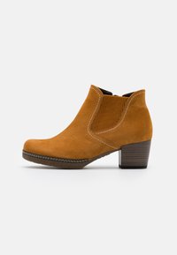 Botas de tobillo de ante en tono marrón claro con un panel lateral elástico texturizado, punta redondeada y un tacón de madera marrón. La costura blanca acentúa el diseño.