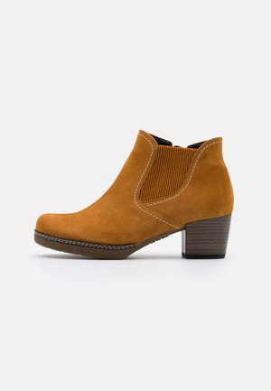 Bottines chelsea en suède tan avec un panneau latéral élastique texturé, un bout arrondi et un talon empilé marron. Des coutures blanches accentuent le design.