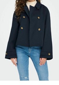 Chaqueta corta de doble botonadura en azul marino con grandes botones dorados, mangas amplias y una textura suave, combinada con jeans claros desgastados.