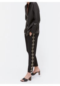 Persoon in zwarte broek met verticale bloemenversieringen aan de zijkant, zwart leren langemouwen shirt en zwarte hoge hakken sandalen.