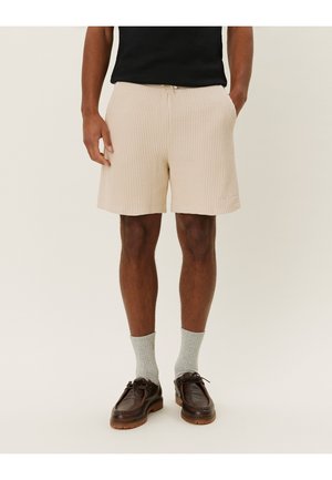 Beige Shorts aus Waffelstruktur mit elastischem Bund, seitlichen Taschen und lockerer Passform, kombiniert mit grauen Socken und braunen Schuhen.