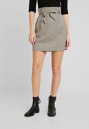 Minifalda de cuadros en beige y negro con una cinturilla alta, detalle de lazo y bolsillos frontales, combinada con botas negras hasta el tobillo.