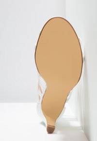 Semelle d'une sandale beige à talon haut avec un petit talon, inclinée contre un mur et une surface blanche.