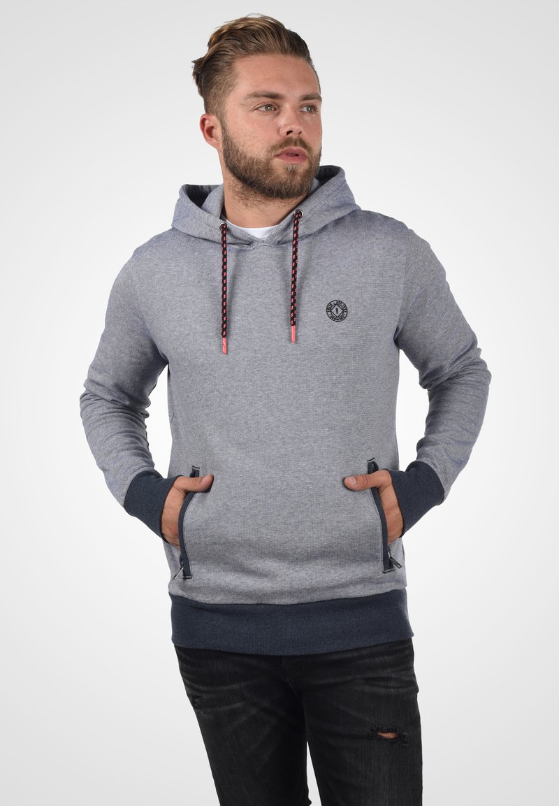 Grauer Kapuzenpullover mit strukturierter Oberfläche, ausgestattet mit einem Logo auf der Brust, kontrastierenden dunklen Bündchen und schwarzen Kordeln mit roten Akzenten.