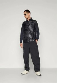 Čierny prešívaný vest s prešívaným dizajnom, skombinovaný s tmavými cargo nohavicami a svetlými teniskami. Doplnky zahŕňajú svetlé slnečné okuliare.