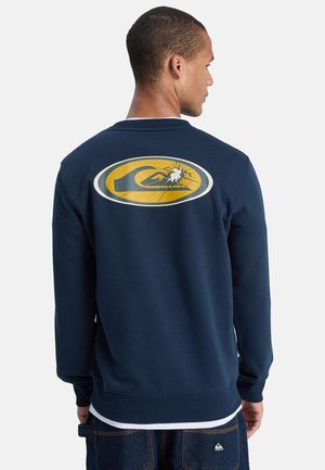 Man die met de rug naar de kijker staat, draagt een marineblauwe trui met een groot, geel-wit gebarsten logo op de achterkant en blauwe jeans met een logopatch.