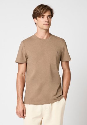Homme debout, une main dans la poche, portant un t-shirt marron à manches courtes et un pantalon de couleur claire sur un fond uni.