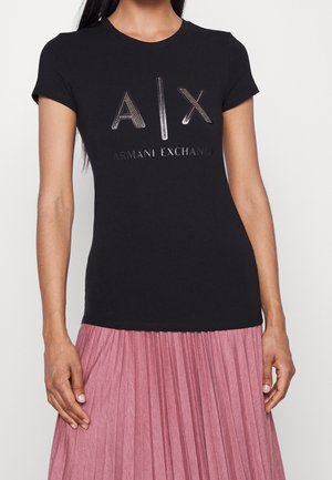 Kobieta ubrana w czarny T-shirt Armani Exchange z ćwiekowanym logo "A|X" i różową plisowaną spódnicę na jasnym jednolitym tle.