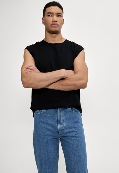 Joven con cabello rizado corto vestido con una camiseta sin mangas negra y jeans azules, de pie con los brazos cruzados frente a un fondo liso.