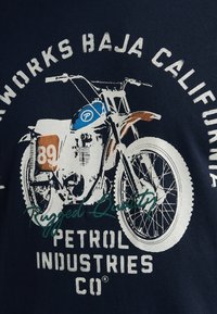 T-shirt bleu marine avec un graphisme de moto vintage en blanc et marron, avec le numéro 89, des accents bleus et un texte stylisé.