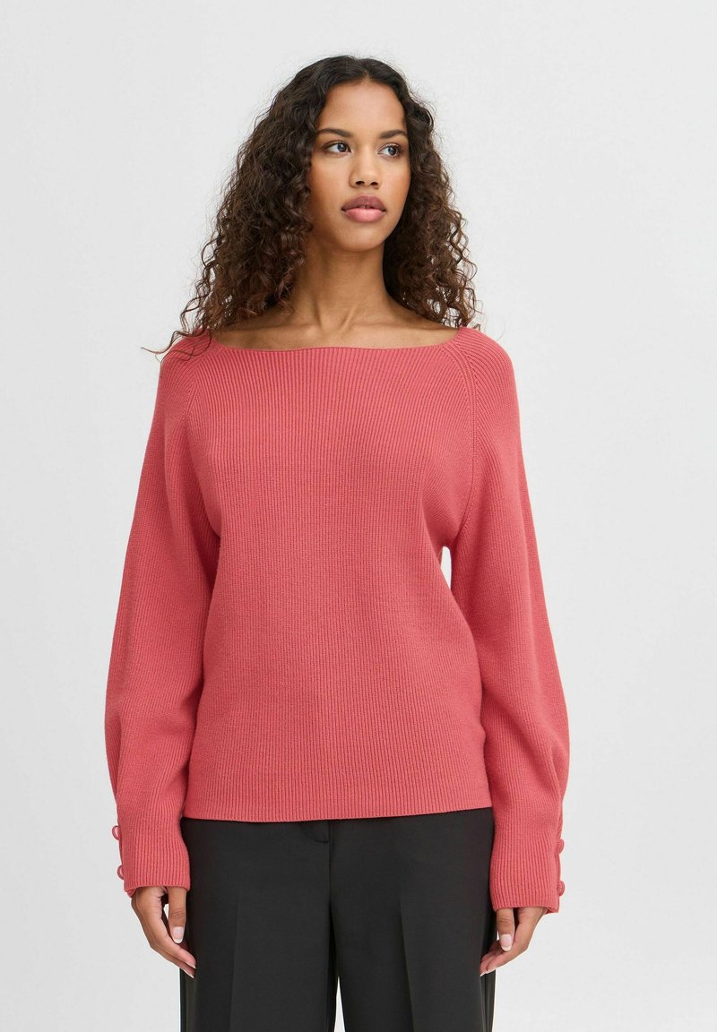 Ribana pulover v koralni barvi z izrezom, ki odkriva ramena, balonastimi rokavi in gumbi na manšetah, v kombinaciji s temnimi hlačami.