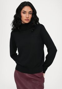 Pull over en col roulé noir en texture côtelée avec des manches longues. Présente un logo subtil en fil sombre. Associé à une jupe en cuir bordeaux.