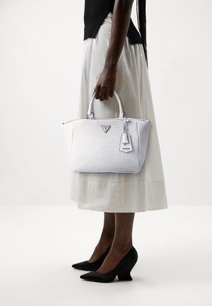 Personne portant une jupe blanche, un haut noir et des talons noirs, tenant un sac à main Guess tissé blanc par ses poignées.