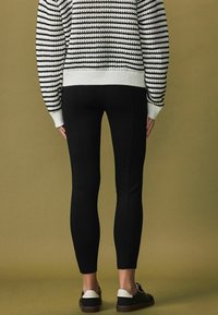 Schwarze Leggings mit glatter Textur, kombiniert mit einem schwarz-weißen gestreiften Strickpullover. Am unteren Rand sind legere Schuhe sichtbar.